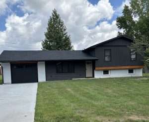 A modern split-level house with dark gray and white siding, a single-car garage, large front windows, and a wide green lawn under a partly cloudy sky.