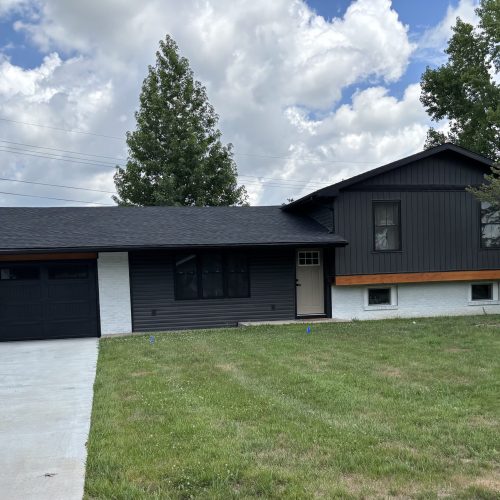 A modern split-level house with dark gray and white siding, a single-car garage, large front windows, and a wide green lawn under a partly cloudy sky.