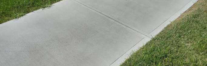 A clean, gray concrete sidewalk runs through a grassy area. The grass on one side is green, while the other side shows patches of dry, brown grass. The sidewalk appears smooth and well-maintained.